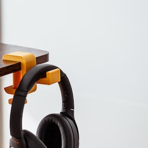 Peut inclure: Un casque audio noir suspendu à une pince de bureau jaune et orange, fixée à un bureau brun foncé.