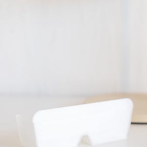 Może przedstawiać: Okulary przeciwsłoneczne z białego plastiku o zaokrąglonym, prostokątnym kształcie. Okulary leżą na białej powierzchni.