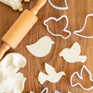 May include: A wooden rolling pin and white play dough on a wooden surface. There are several white plastic bird-shaped cookie cutters.