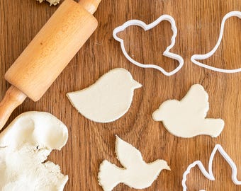Peut inclure: Un rouleau à pâtisserie en bois et de la pâte à modeler blanche sur une surface en bois. Il y a plusieurs emporte-pièces en forme d'oiseau en plastique blanc.