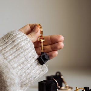May include: A hand holding a gold key chain with a black camera charm. Three black camera charms are on a white surface.