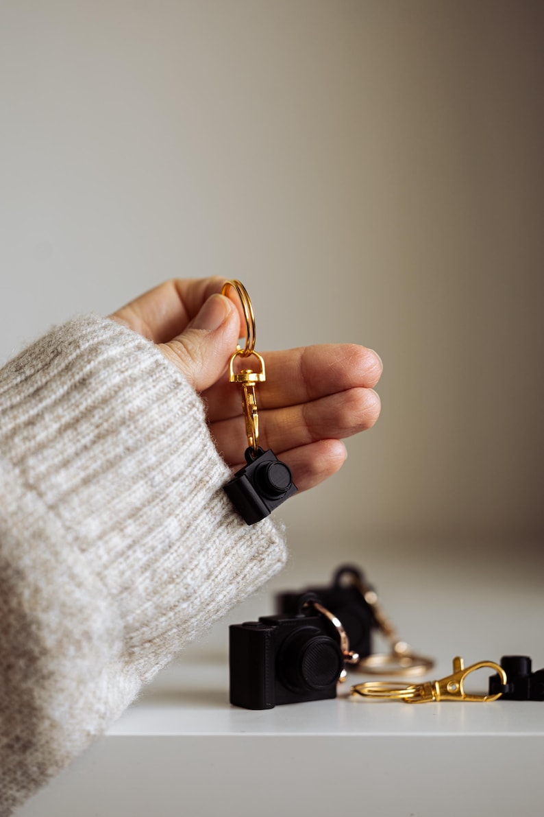May include: A hand holding a gold key chain with a black camera charm. Three black camera charms are on a white surface.