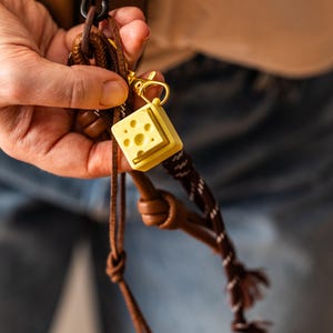 以下が含まれることがあります： 茶色の革製のバッグに、金色と黄色のチーズのキーホルダーが付いています。キーホルダーは柔らかく、弾力のある素材でできており、リアルなチーズの質感があります。