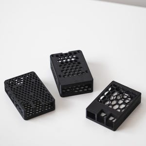 Op de afbeelding: Drie zwarte rechthoekige behuizingen met een honingraatpatroon. De behuizingen zijn gemaakt van een matzwart materiaal en hebben een geometrisch ontwerp. De behuizingen zijn op een wit oppervlak geplaatst.