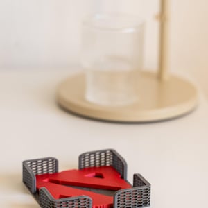 Puede incluir: Un posavasos de plástico gris con un logotipo rojo en forma de una letra estilizada "N" en su interior. El posavasos está diseñado para sostener una taza o un vaso.