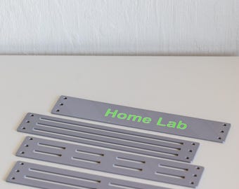 May include: A set of five rectangular grey panels made of a smooth material. The top panel features the text "Home Lab" in bright green lettering. Each panel has three small circular holes on both the left and right ends for mounting purposes.