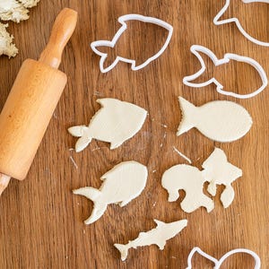 May include: A wooden rolling pin and white plastic cookie cutters shaped like fish and a seahorse on a wooden surface. The cookie cutters are used to cut out shapes from play dough.