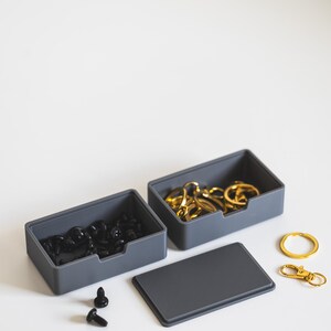 May include: Two grey rectangular storage containers. One contains black hardware, the other gold keyrings. A grey lid is nearby. Two gold keychains and a key ring are also visible.