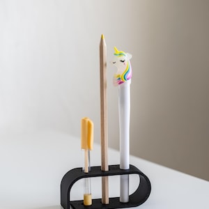 Puede incluir: Un organizador de escritorio negro contiene un l&aacute;piz amarillo, un bol&iacute;grafo transparente con tapa amarilla y un bol&iacute;grafo blanco coronado con una cabeza de unicornio. El organizador est&aacute; sobre una superficie blanca.