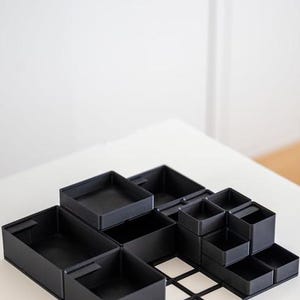 Op de afbeelding: Een modulaire zwarte bureau-organizer met een rasterbasis en meerdere vierkante en rechthoekige vakken van verschillende hoogtes. Het systeem bevat diverse open bakjes die op de platte basis met open frame kunnen worden geplaatst.