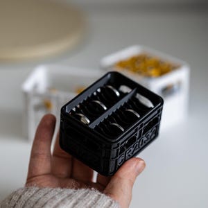 Op de afbeelding: Een zwarte plastic krat met een grijze scheidingswand, met daarin verschillende zilveren ringen. Op de zijkant van de krat staat "GRAUS" gedrukt.