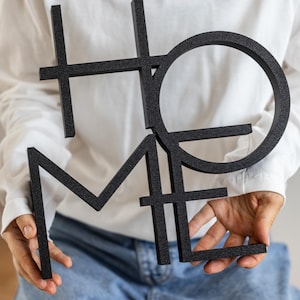 May include: A black, three-dimensional wall sign featuring the word HOME in a stylized, interconnected block font. The letters are arranged in a stacked, geometric layout with a textured, matte finish, creating a bold and modern visual statement.