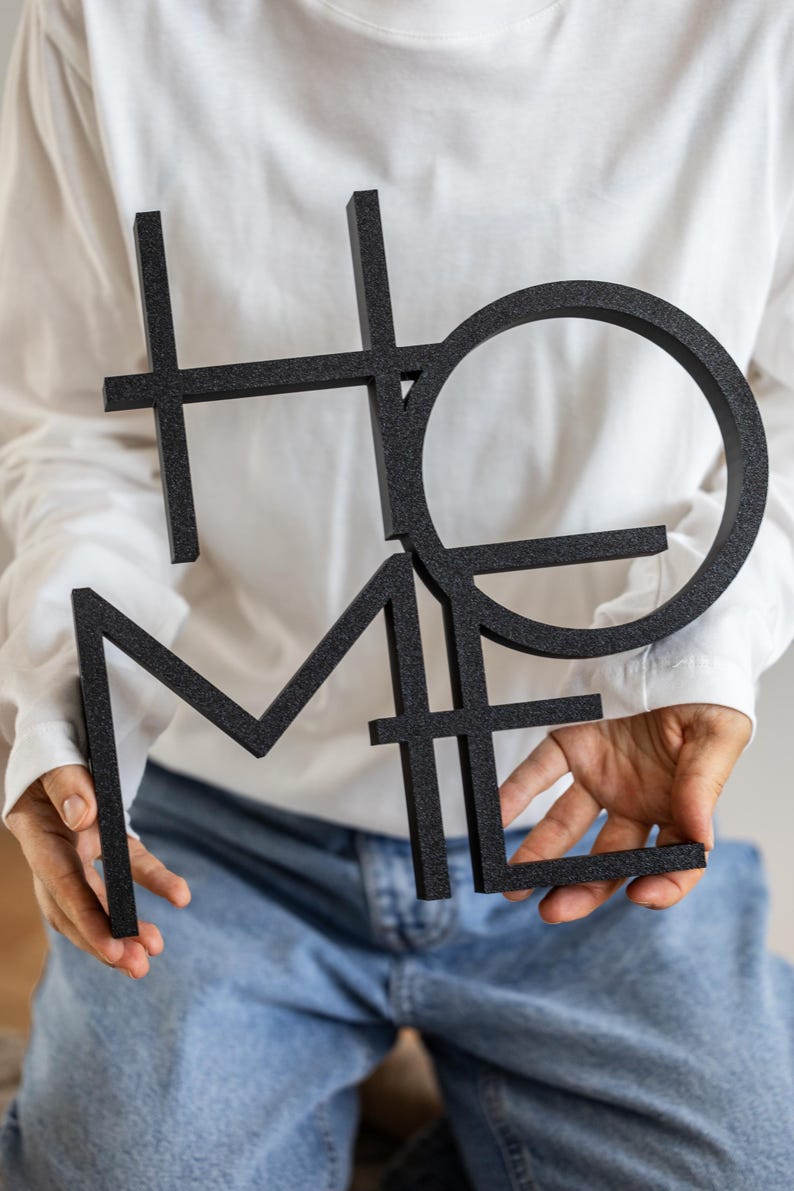 May include: A black, three-dimensional wall sign featuring the word HOME in a stylized, interconnected block font. The letters are arranged in a stacked, geometric layout with a textured, matte finish, creating a bold and modern visual statement.