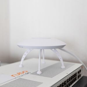 Pode incluir: Um ponto de acesso Aruba Instant On branco, que se assemelha a um pequeno dispositivo futurista, está em cima de um switch de rede. O ponto de acesso tem um design circular com três pernas brancas. O switch é branco com texto laranja.