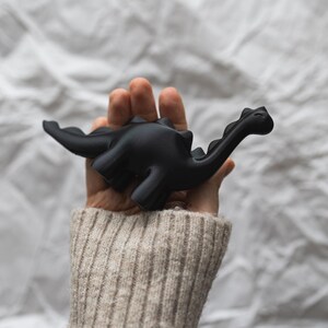 May include: A hand holding a black dinosaur toy. The dinosaur has a long neck and tail.