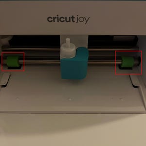 Pode incluir: Uma máquina de corte Cricut Joy branca e azul-turquesa com um carro central e dois rolos de borracha verdes posicionados numa haste metálica horizontal. A máquina tem uma base branca e uma tampa superior turquesa com o texto Cricut Joy.