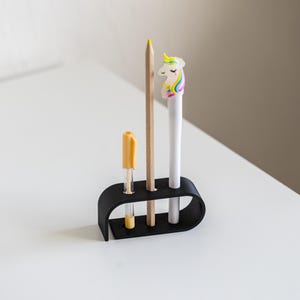 Puede incluir: Un organizador de escritorio negro que contiene un bol&iacute;grafo amarillo, un l&aacute;piz de madera y un bol&iacute;grafo blanco con una figura de unicornio. El organizador est&aacute; sobre una superficie blanca.