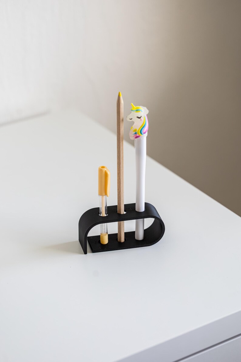 Puede incluir: Un organizador de escritorio negro que contiene un bol&iacute;grafo amarillo, un l&aacute;piz de madera y un bol&iacute;grafo blanco con una figura de unicornio. El organizador est&aacute; sobre una superficie blanca.