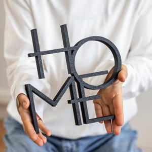 May include: A black, glittery, three-dimensional sign featuring the word HOME arranged in a stacked, block-letter design. The letters are interconnected, with the H and O positioned above the M and E, creating a singular, cohesive decorative object.
