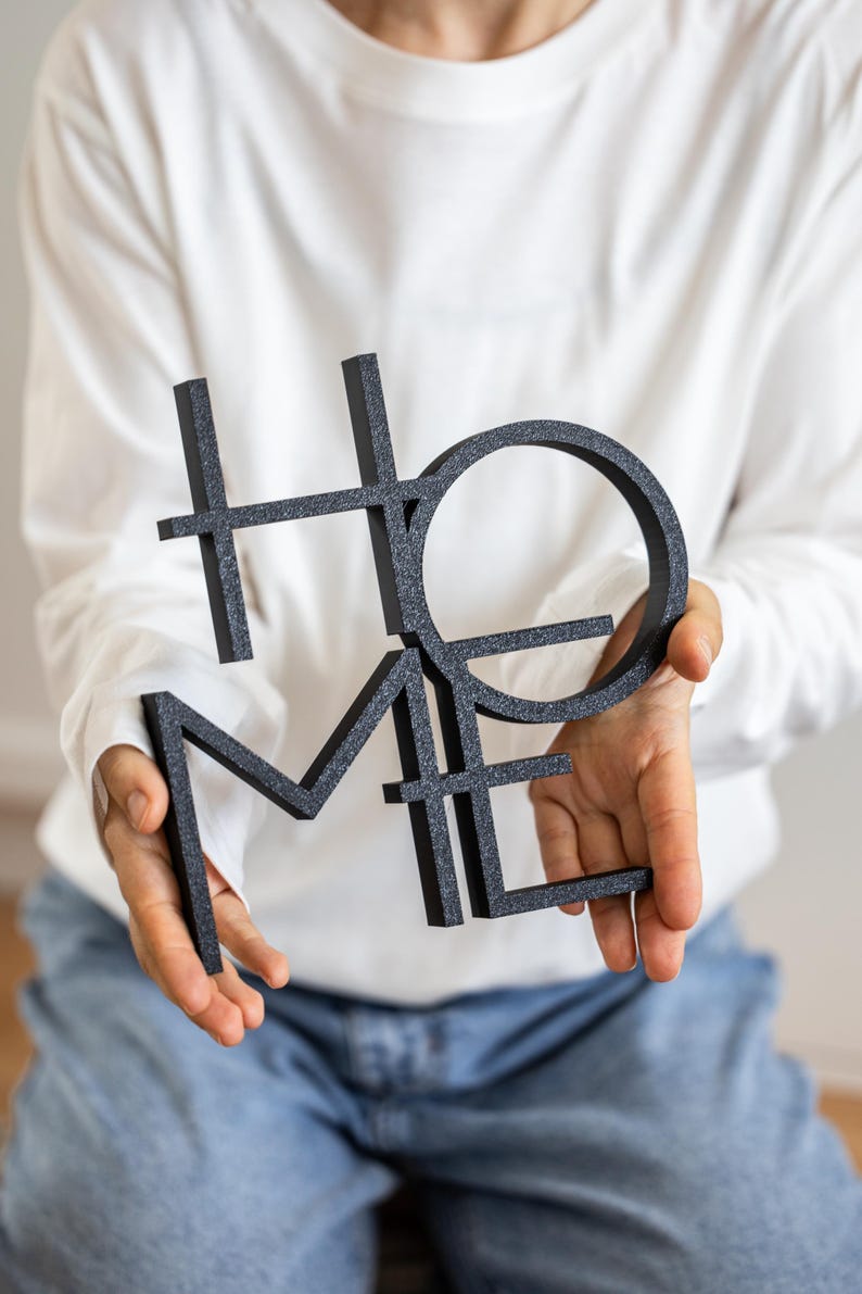 May include: A black, glittery, three-dimensional sign featuring the word HOME arranged in a stacked, block-letter design. The letters are interconnected, with the H and O positioned above the M and E, creating a singular, cohesive decorative object.