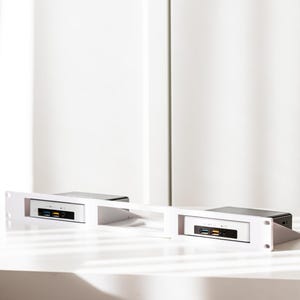 May include: Two small, black and silver computer units are mounted in a white rack. The rack is on a white surface, with a white background. Each unit has USB ports and a power button.