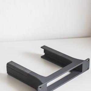 Może przedstawiać: Ciemnoszary wspornik w kształcie litery U o teksturowanej powierzchni. Wspornik posiada prostokątny otwór i dwa otwory montażowe z jednej strony. Prawdopodobnie element elektroniczny lub meblowy.