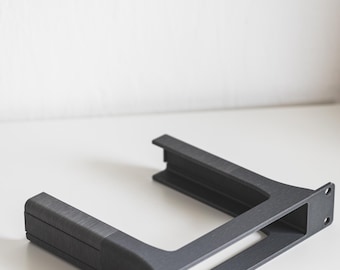 May include: A dark grey, U-shaped bracket with a textured surface. The bracket has a rectangular opening and two mounting holes on one side. Likely a component for electronics or furniture.
