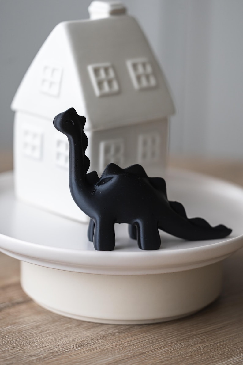 May include: A black dinosaur figurine on a white cake stand. The dinosaur has a long neck and tail and is standing on all fours.