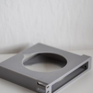 Può includere: Una staffa di montaggio a rack quadrata di colore grigio, dotata di un'ampia apertura circolare al centro e di un pannello frontale piatto con tre fori di montaggio verticali sul lato, progettata per fissare apparecchiature in un rack.