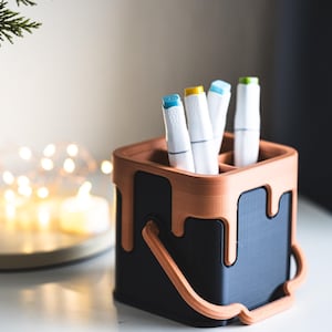 Puede incluir: Un organizador de escritorio en forma de lata de pintura negra y marrón con asa. El organizador tiene cuatro ranuras para bolígrafos o marcadores. Hay cuatro marcadores blancos con tapas de colores en el organizador.