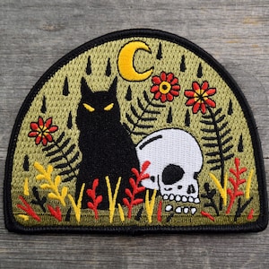 May include: A black cat with yellow eyes sits next to a white skull with a black eye socket. The image is embroidered on a green patch with yellow flowers and a yellow crescent moon.