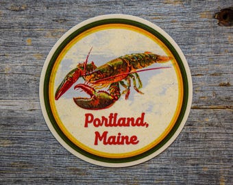 Op de afbeelding: Een ronde sticker met een gedetailleerde illustratie van een kreeft in rood-, oranje- en groentinten. De tekst Portland, Maine staat in dikke rode letters onder de kreeft gedrukt, alles omgeven door een gele, oranje en groene rand.