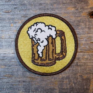 May include: A brown and white embroidered patch of a beer mug with foam on top, on a yellow background.