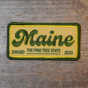 May include: A rectangular patch with a tan background and a green border. The word "Maine" is embroidered in large, cursive green letters. Below, it reads "DIRIGO THE PINE TREE STATE 1820" in smaller green text.