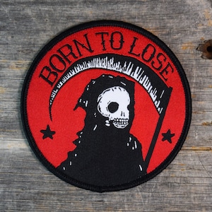 May include: A black and red embroidered patch with a white skull and scythe. The patch reads "Born to Lose" in a black circular border.