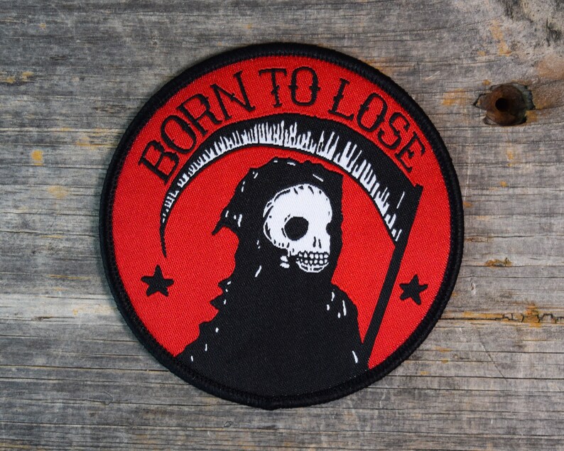 Op de afbeelding: Een geborduurde zwarte en rode patch met een witte schedel en een zeis. De patch heeft de tekst "Born to Lose" in een zwarte cirkel.