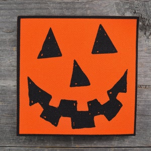 May include: Orange canvas with a black painted jack-o'-lantern face. The face has two triangle eyes, a triangle nose, and a wide, toothy grin.