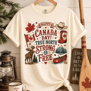 May include: Cream-coloured t-shirt with a graphic celebrating Canada Day. The design includes a maple leaf, Canadian flag, cowboy hat, boots, moose, canoe, and the text "Happy Canada Day! True North Strong & Free."