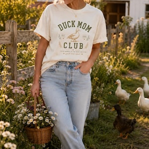 May include: Cream-colored t-shirt with the text "DUCK MOM CLUB" and a duck illustration. The shirt is paired with light blue jeans and white sneakers. A woman is holding a basket of flowers in a garden setting with ducks.