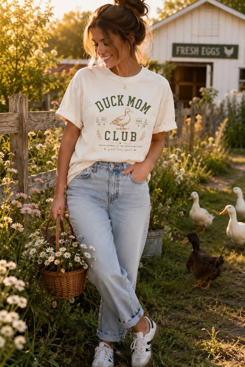 May include: Cream-colored t-shirt with the text "DUCK MOM CLUB" and a duck illustration. The shirt is paired with light blue jeans and white sneakers. A woman is holding a basket of flowers in a garden setting with ducks.