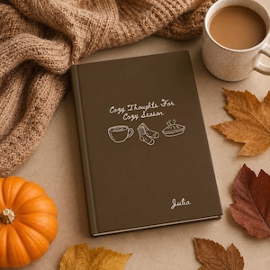 Puede incluir: Un diario de tapa dura de color marrón oscuro con el texto blanco "Cozy Thoughts For Cozy Season" sobre ilustraciones de línea de una taza de café, calcetines y un pastel, con el nombre "Julia" impreso en blanco en la esquina inferior derecha.