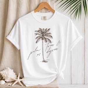 May include: A white short-sleeve t-shirt featuring a dark brown graphic of a single palm tree centred on the chest. The text "palm trees & 90 degrees" is written in a cursive script font across the graphic. The shirt has a crew neckline.