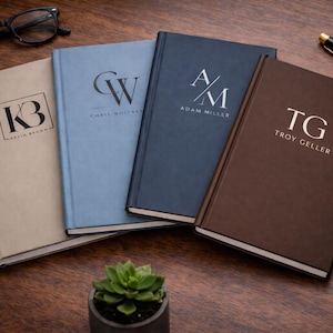 May include: Four personalized notebooks in beige, light blue, navy blue, and brown. Each notebook features initials and names: KB, Chris Whitak, AM, and Troy Geller. A pair of glasses, a pen, and a small succulent are also visible.