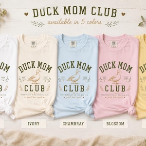 May include: Five t-shirts in white, ivory, chambray, blossom, and butter colors are displayed. Each shirt features the text "DUCK MOM CLUB" with a duck illustration. The text "available in 5 colors" is above the shirts.