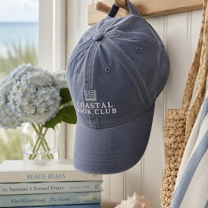 May include: A slate blue baseball cap with a white embroidered open book icon and the text COASTAL BOOK CLUB centred on the front panel. The cap features a structured crown, a curved brim, and a fabric strap with a metal buckle at the back.