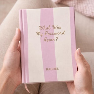May include: A pink and white striped journal with the text "What Was My Password Again?" in gold lettering. The name "RACHEL" is printed at the bottom. The journal is held open, revealing a pink ribbon bookmark.