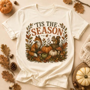 May include: A cream-colored short-sleeve t-shirt featuring a graphic print with the text 'TIS THE SEASON' in dark green and orange. The design includes a fox, a squirrel, pumpkins, mushrooms, and autumn leaves in shades of orange and brown.