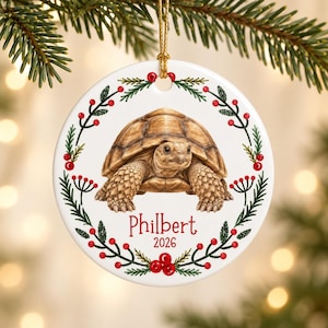 May include: A round white ceramic ornament featuring a detailed illustration of a brown tortoise in the center. A wreath of green pine needles and red berries encircles the tortoise. The name Philbert and the year 2026 are printed in red.