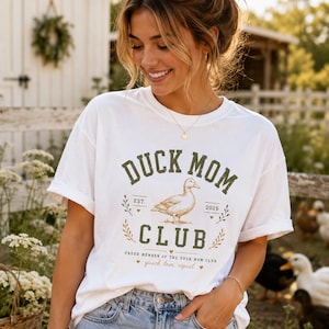 May include: White t-shirt with a green and gold graphic that reads "DUCK MOM CLUB" with a duck illustration. The shirt also has the text "quack, love, repeat". The person is wearing blue jeans.