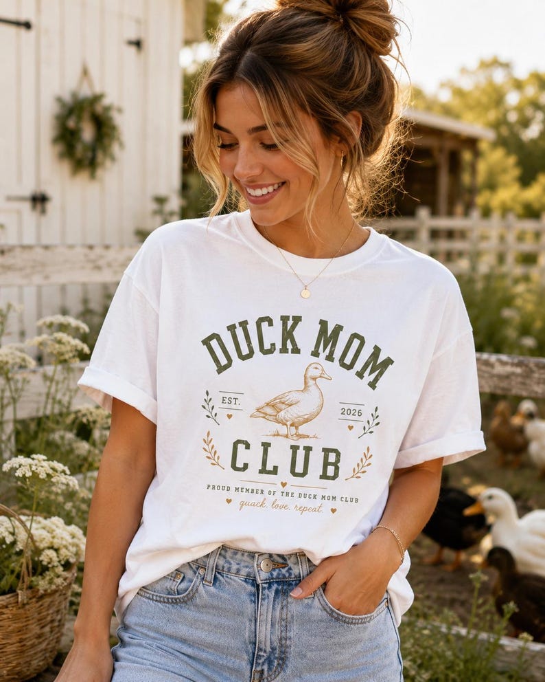 May include: White t-shirt with a green and gold graphic that reads "DUCK MOM CLUB" with a duck illustration. The shirt also has the text "quack, love, repeat". The person is wearing blue jeans.